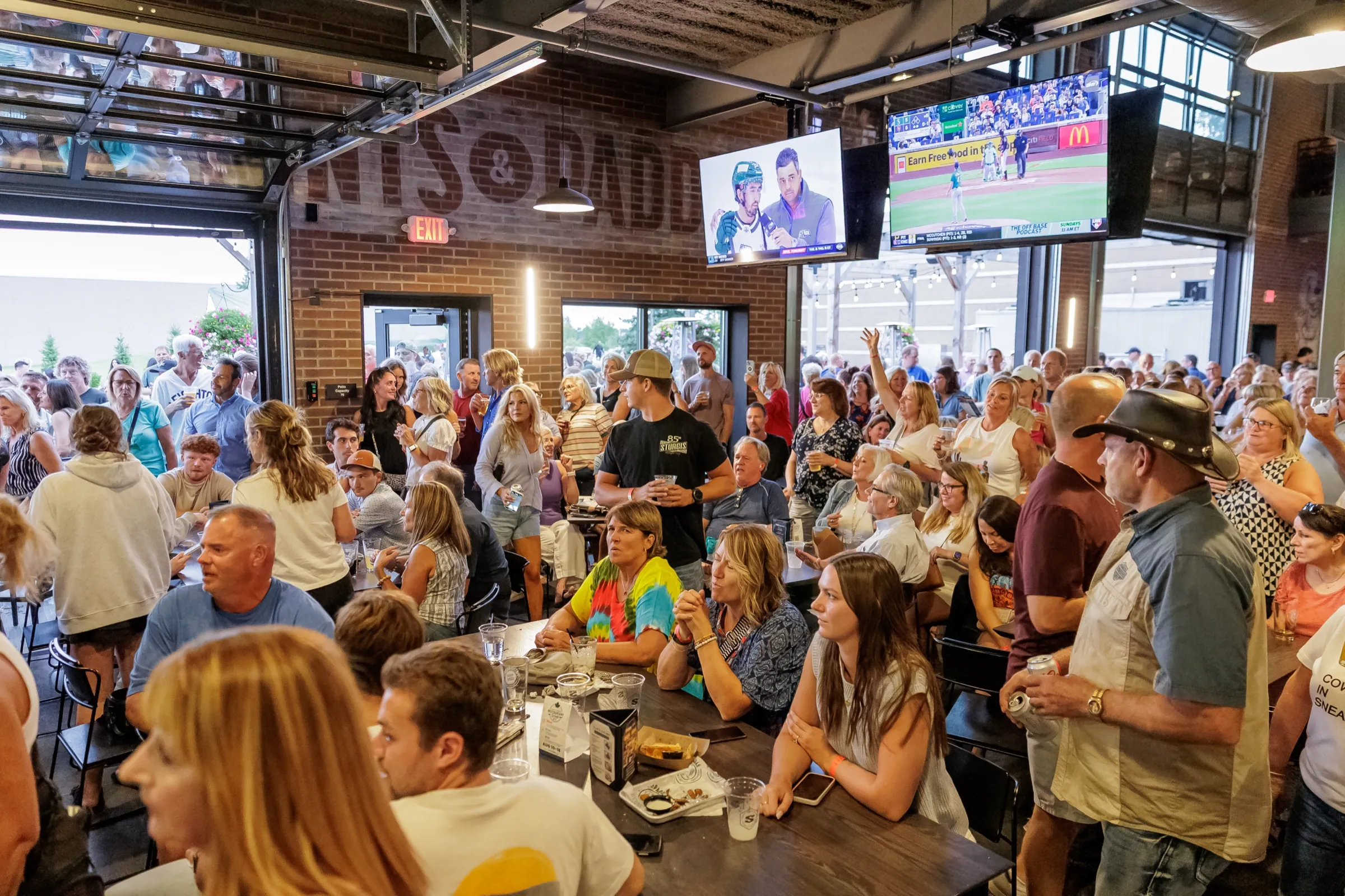 A packed main-level crowd at the long wood tables with TVs overhead and the open garage doors behind