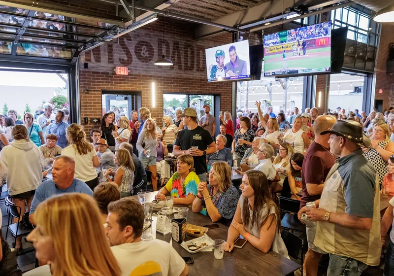 A packed main-level taproom with guests at long wood tables, TVs overhead and the open garage doors behind