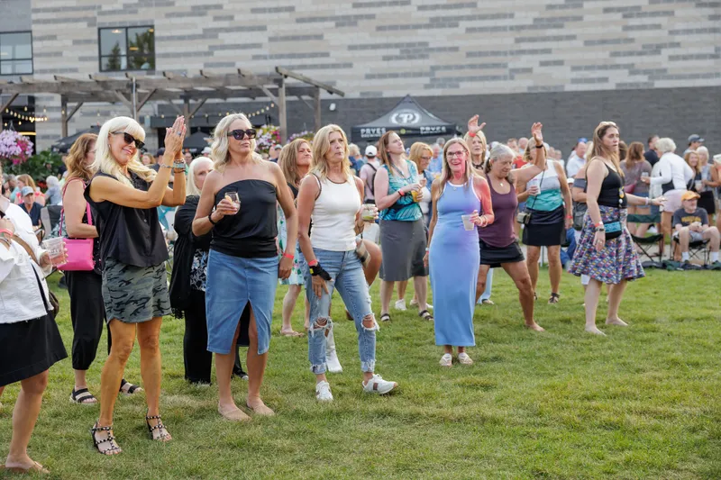 A group of friends dancing and clapping on the lawn, drinks in hand, with the crowd, the stone building and the pergola behind them