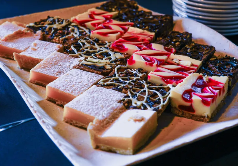 A white tray of assorted dessert bars: lemon, brownie, raspberry cheesecake and chocolate swirl