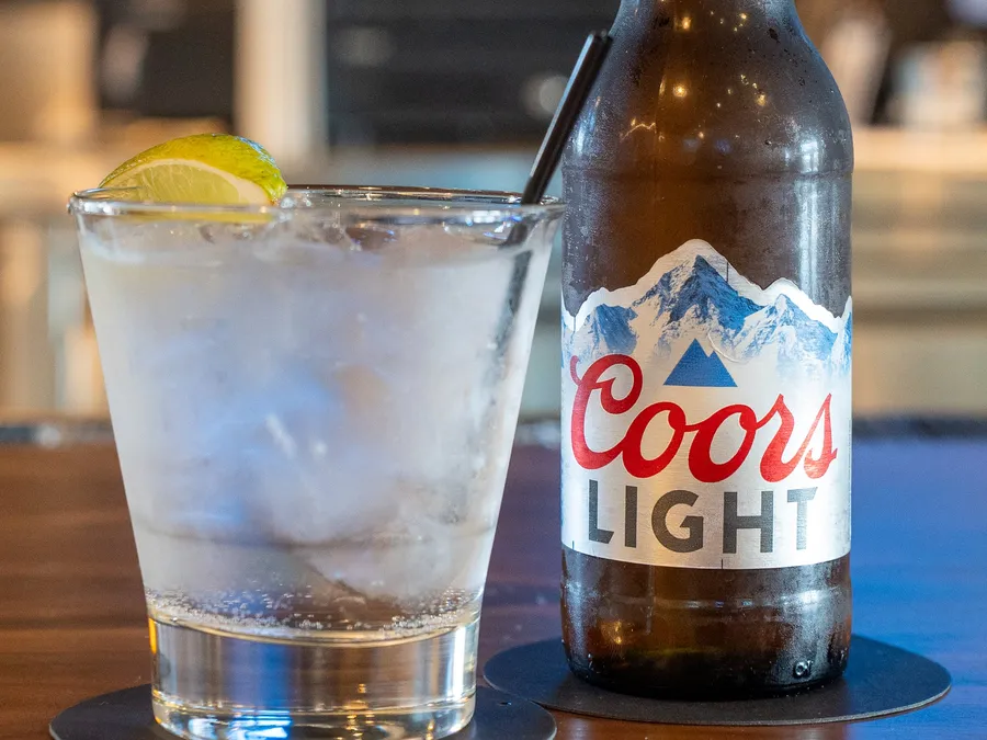 A Coors Light bottle beside a clear rail drink over ice with a lime wedge on the wooden bar