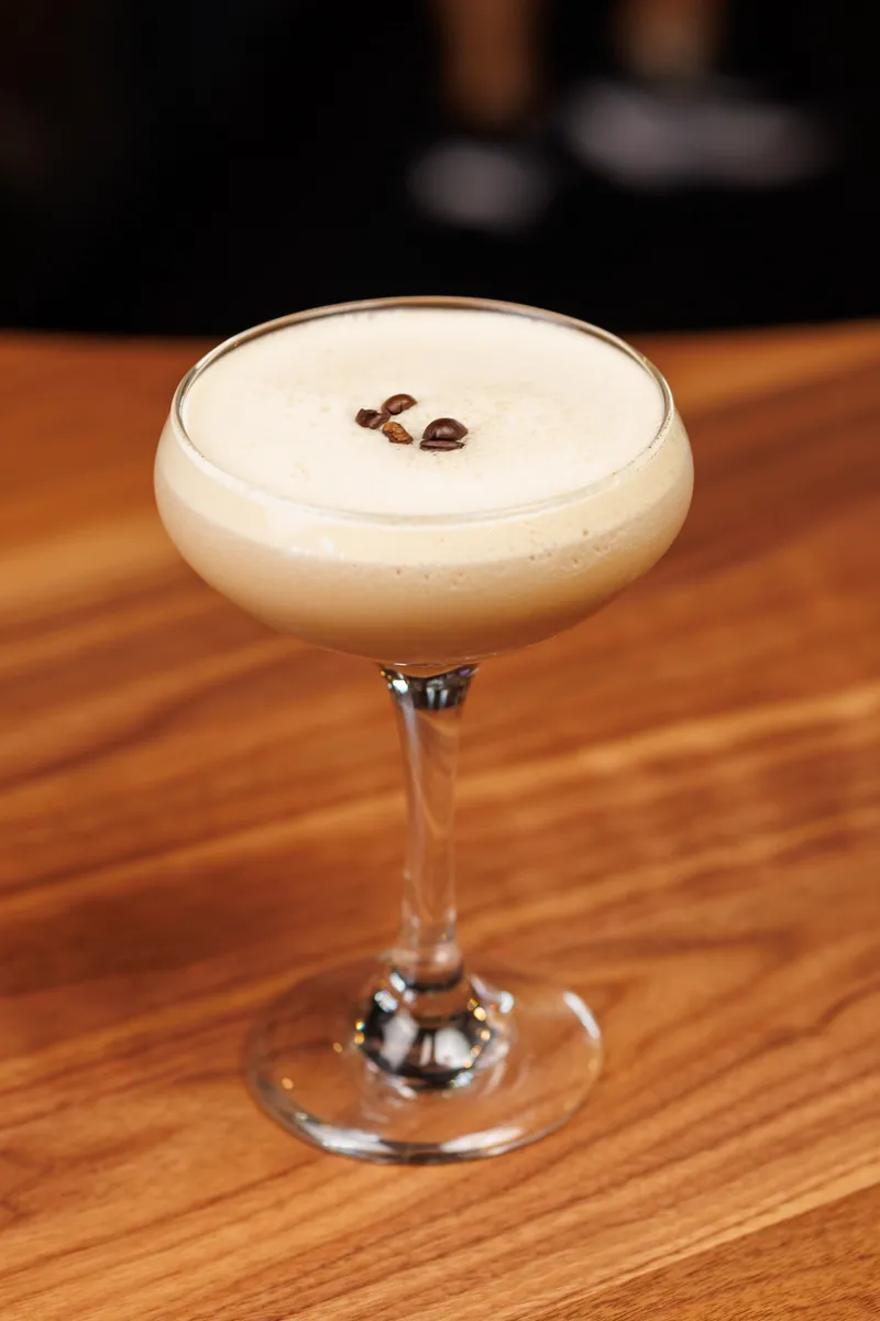 An espresso martini in a coupe with three coffee beans on the foam, on the walnut bar top