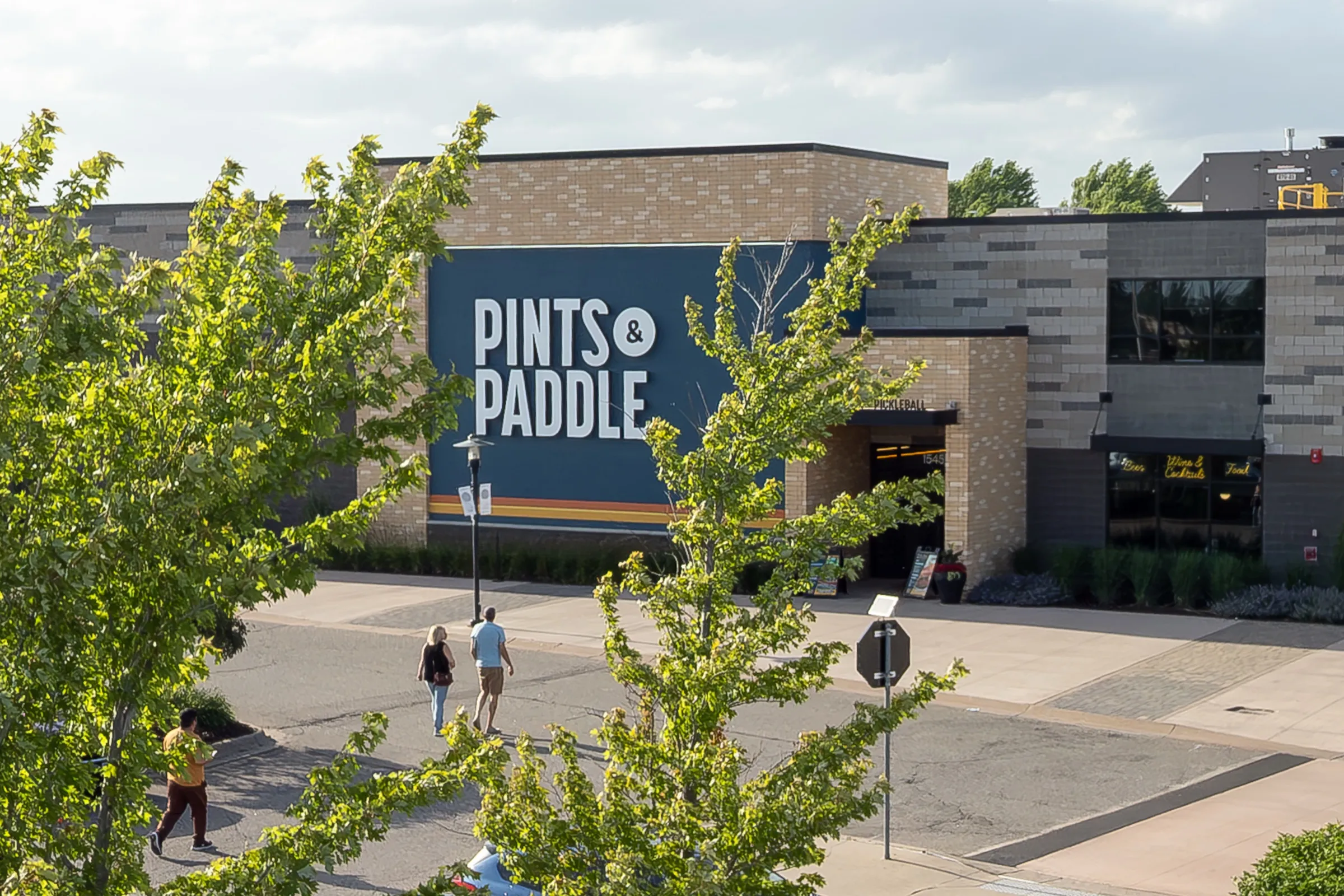 Street-level view of the brick and stone front of the building on a sunny evening, with the navy PINTS & PADDLE sign, the PICKLEBALL entrance and a few people walking toward the door