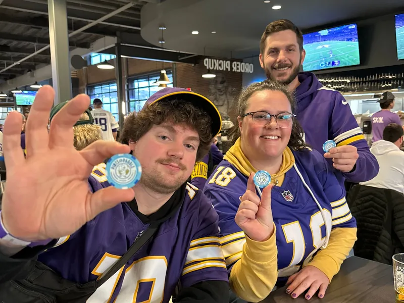 Three friends in Vikings jerseys at a table hold up their light-blue Pints & Paddle drink chips