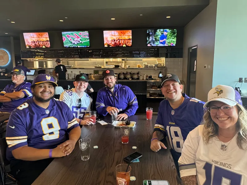 Five friends in Vikings jerseys around a table in front of the kitchen counter, TVs above