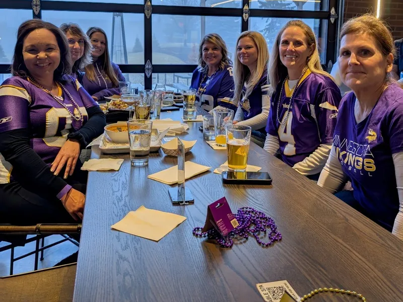 Eight women in Vikings jerseys down both sides of a long wood table, beads and beers in front of them, the garage door behind