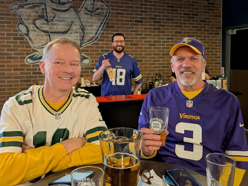 A Packers fan and a Vikings fan in a 3 jersey share a table and a pitcher of beer under the Paddy mural, a bartender in an 18 jersey behind