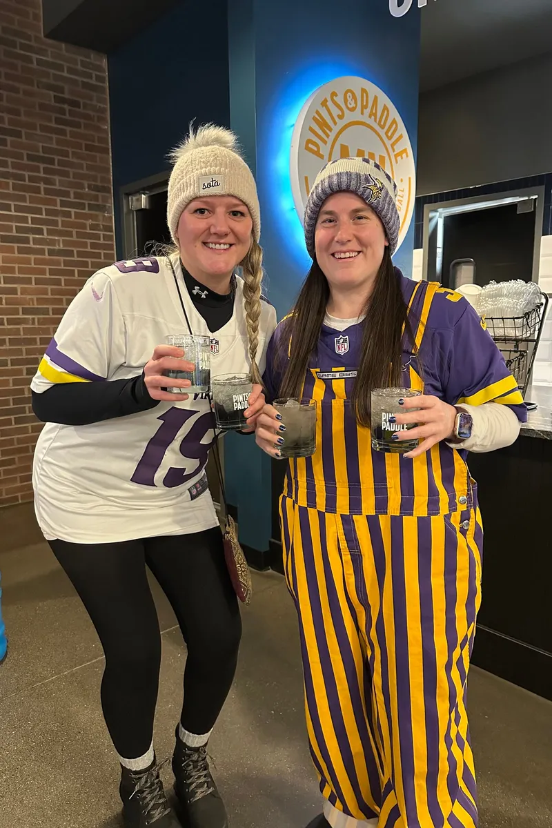 Two fans toast with rocks glasses under the Pints & Paddle sign, one in a white 19 jersey and knit hat, the other in purple-and-gold striped overalls