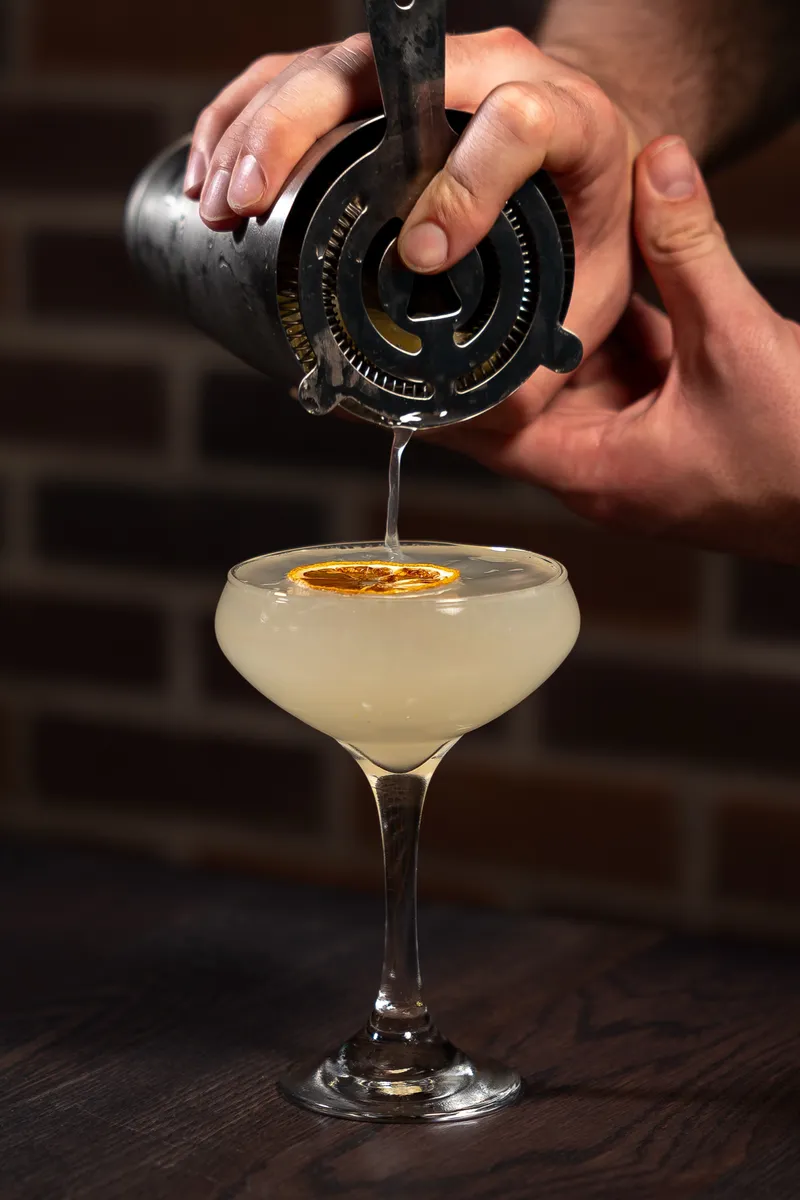 A bartender's hands double-straining a French 75 from a shaker into a coupe garnished with a dried orange wheel