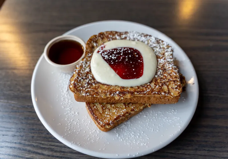Two stacked slices of cinnamon French toast with whipped cream cheese, raspberry sauce and powdered sugar, syrup on the side