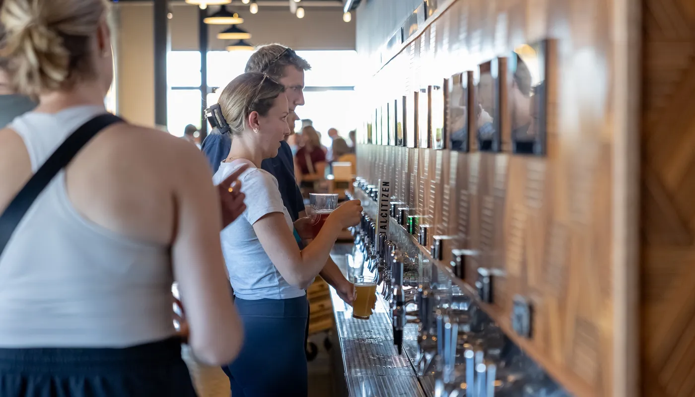 Guests pouring their own beers along the self-serve tap wall, one filling a glass under a row of tap handles