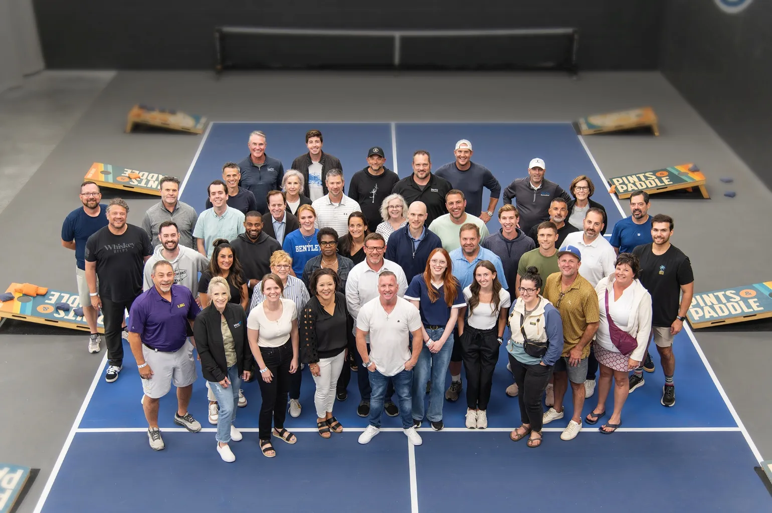 An event group of about forty-five guests smiling up at the camera from a blue indoor court, with Pints & Paddle bags boards set up around them