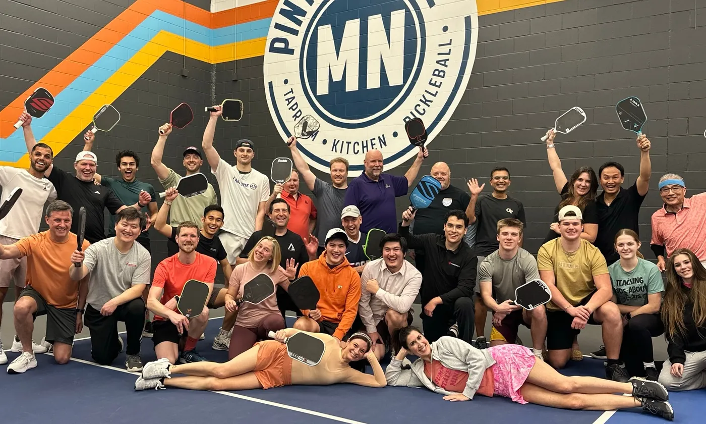 A company group of about thirty posing with paddles raised on a court under the Pints & Paddle MN wall logo