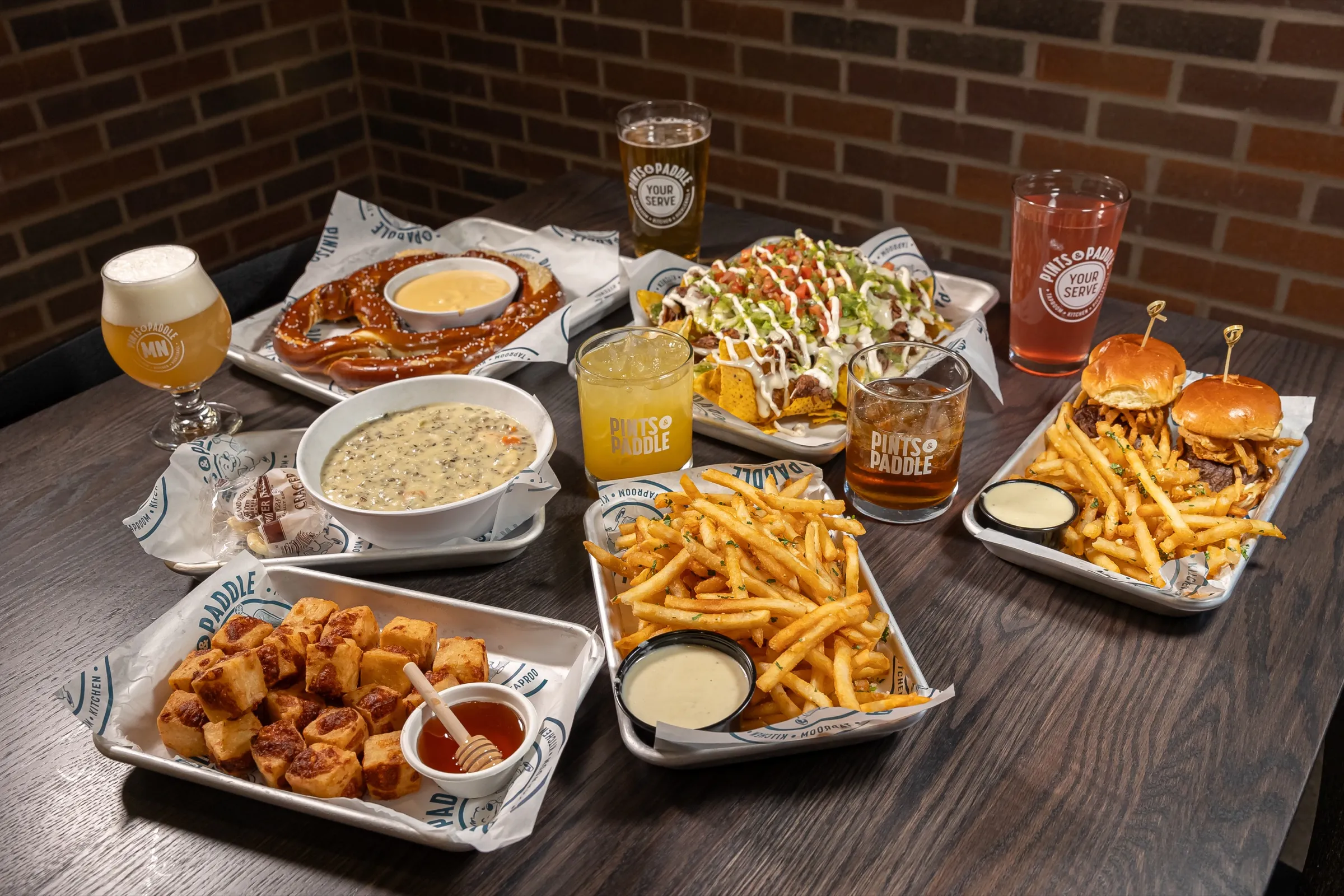 A table spread of pretzel with cheese, loaded nachos, wild rice soup, cheese curds, fries, sliders and four drinks against a brick wall