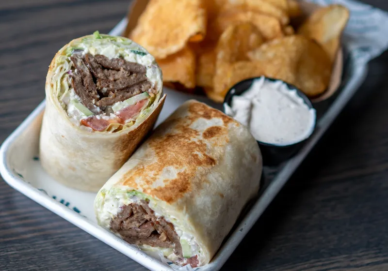 A gyro wrap cut in half showing gyro meat, lettuce, tomato, cucumber, feta and red onion, with kettle chips and a cup of tzatziki