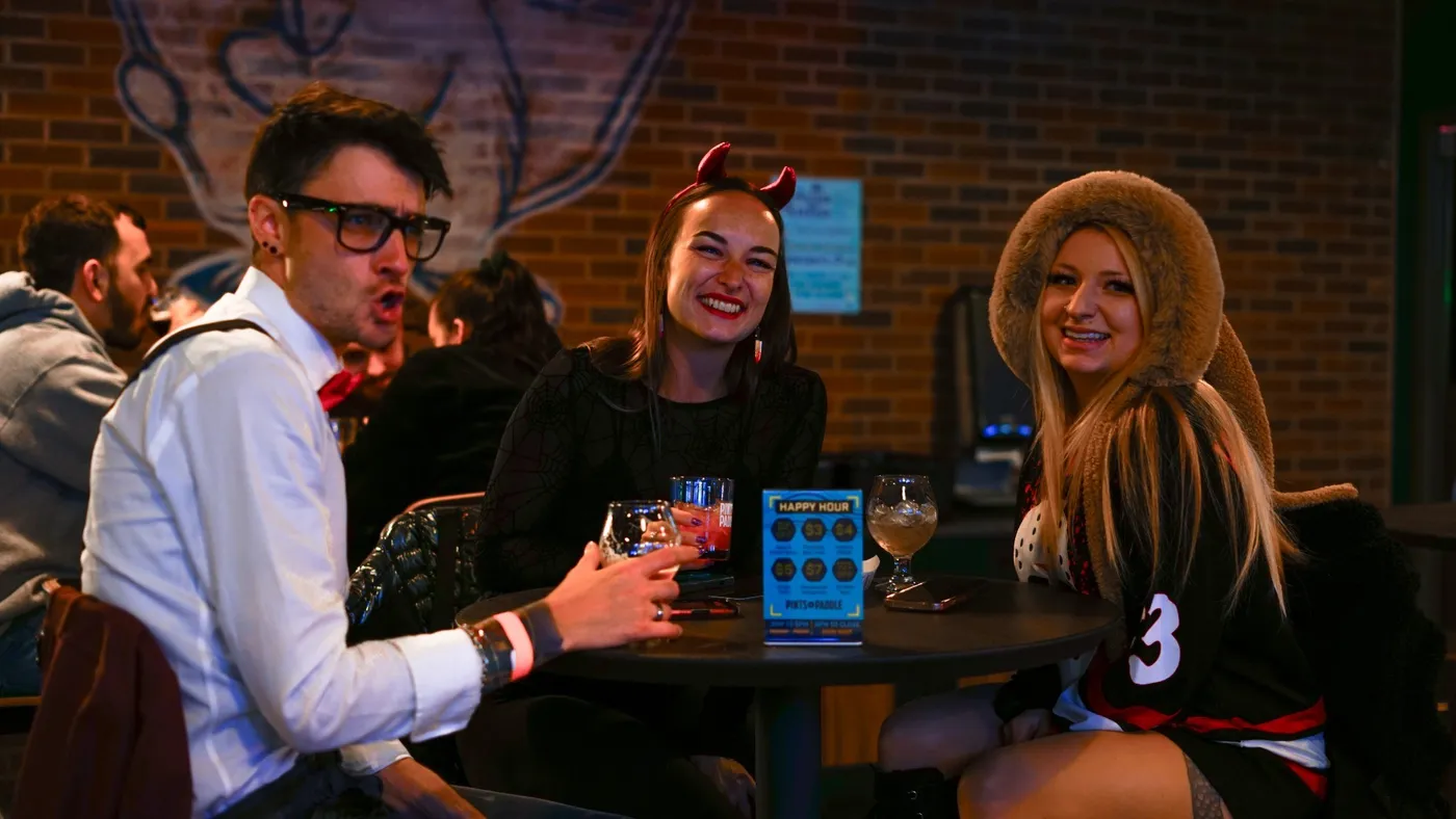 Three friends laughing at a table by the brick wall, one in devil horns, one in a fuzzy hood and one in a bow tie, a happy hour card between their drinks