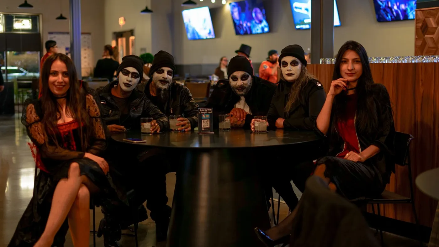 Six friends in ghoulish white face paint, beanies and black leather around a high-top table, drinks in hand, TVs glowing behind them