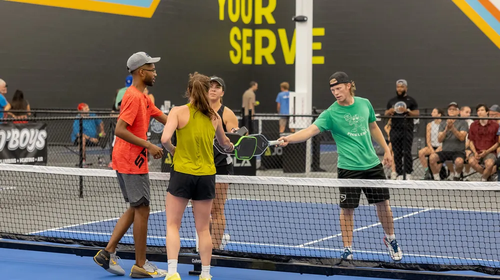 Four players tap paddles at the net after an open play game in front of the Your Serve wall