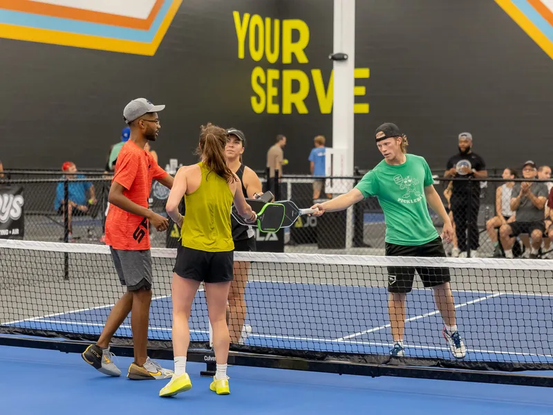 Four players tap paddles at the net in front of the Your Serve wall