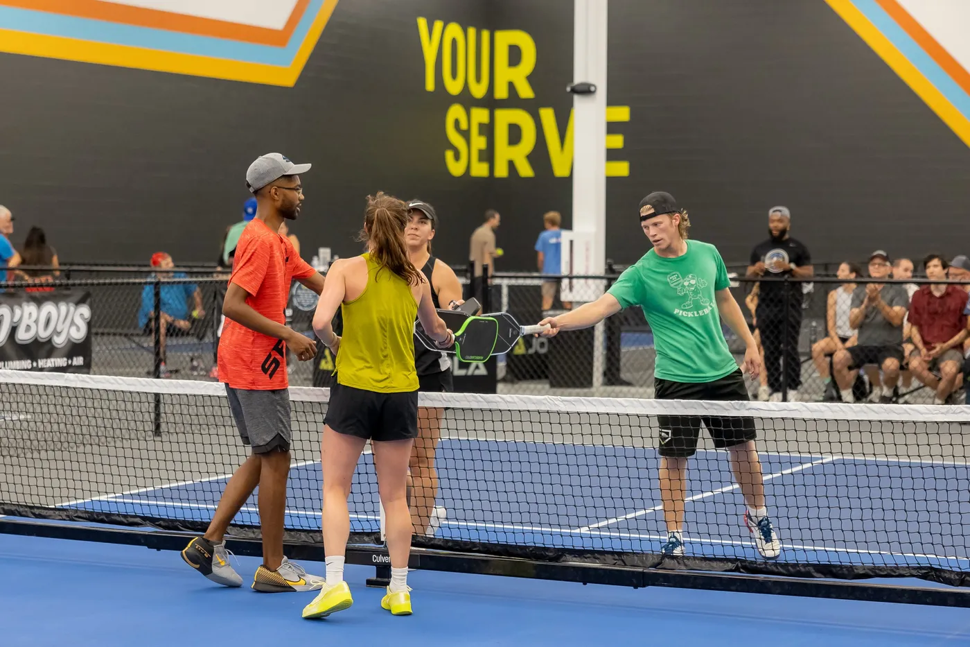Four players tap paddles at the net after a game in front of the yellow Your Serve wall