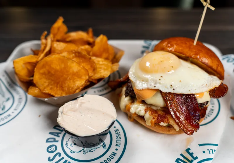 The Hangover Burger on branded paper: a sunny-side-up egg over maple pepper bacon, melted pepper jack and cheese curds on a brioche bun, with house kettle chips and a cup of sauce
