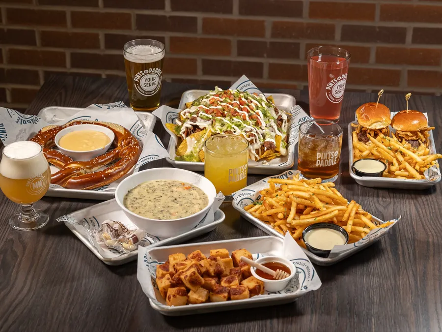 A high-angle spread of pretzel with beer cheese, loaded nachos, sliders with fries, cheese curds, a hazy beer and cocktails
