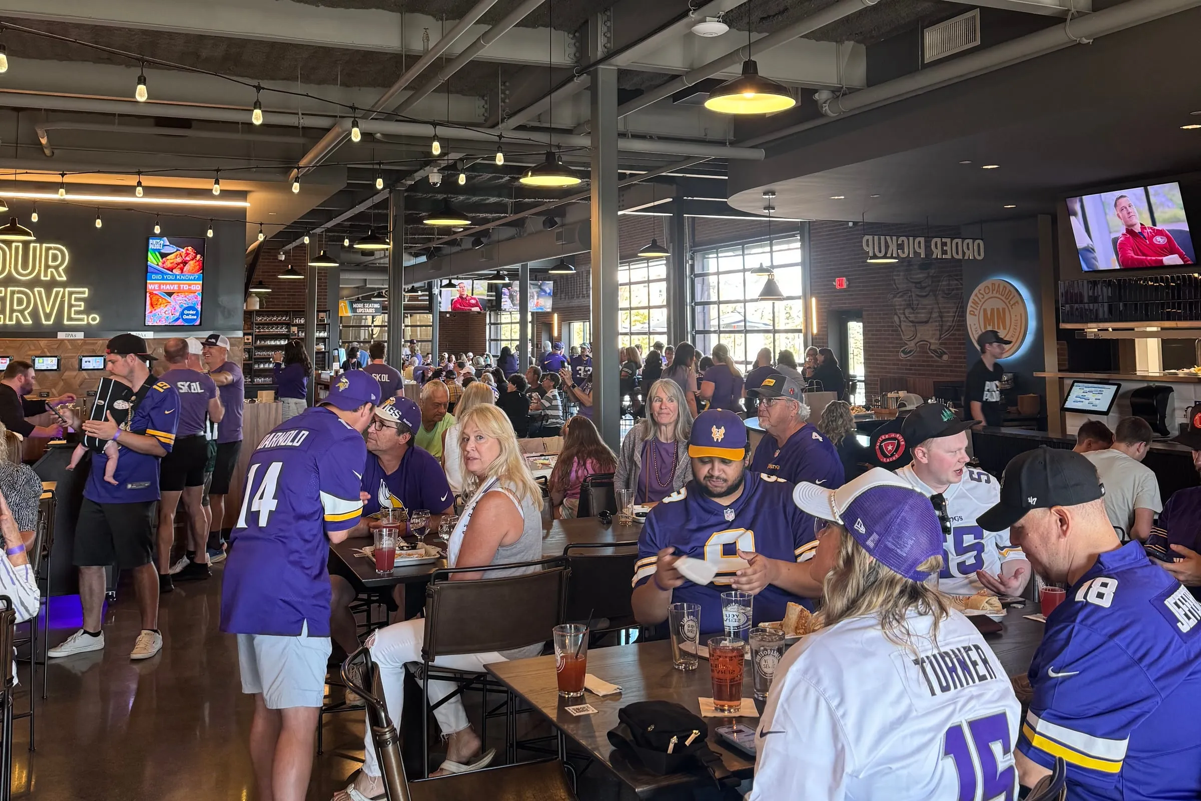 The main level packed with fans in purple Vikings jerseys at the long tables on a Sunday, the YOUR SERVE sign and TVs along the far wall