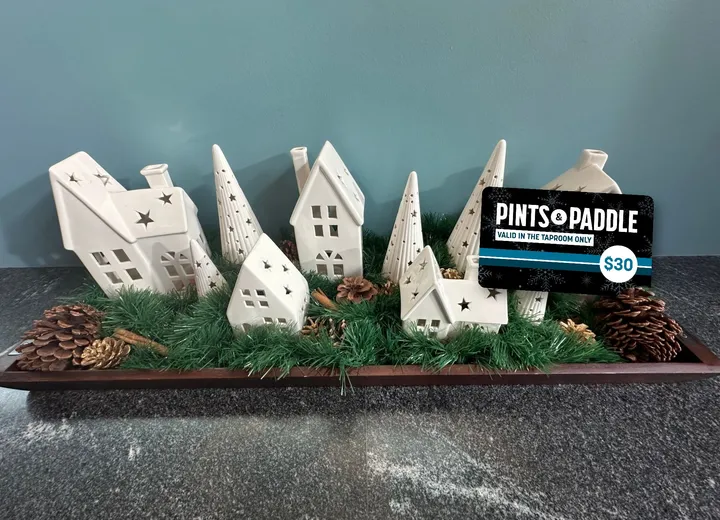 A $30 holiday bonus card propped in the greenery of a white ceramic holiday village, houses and trees with star cut-outs on a wooden tray