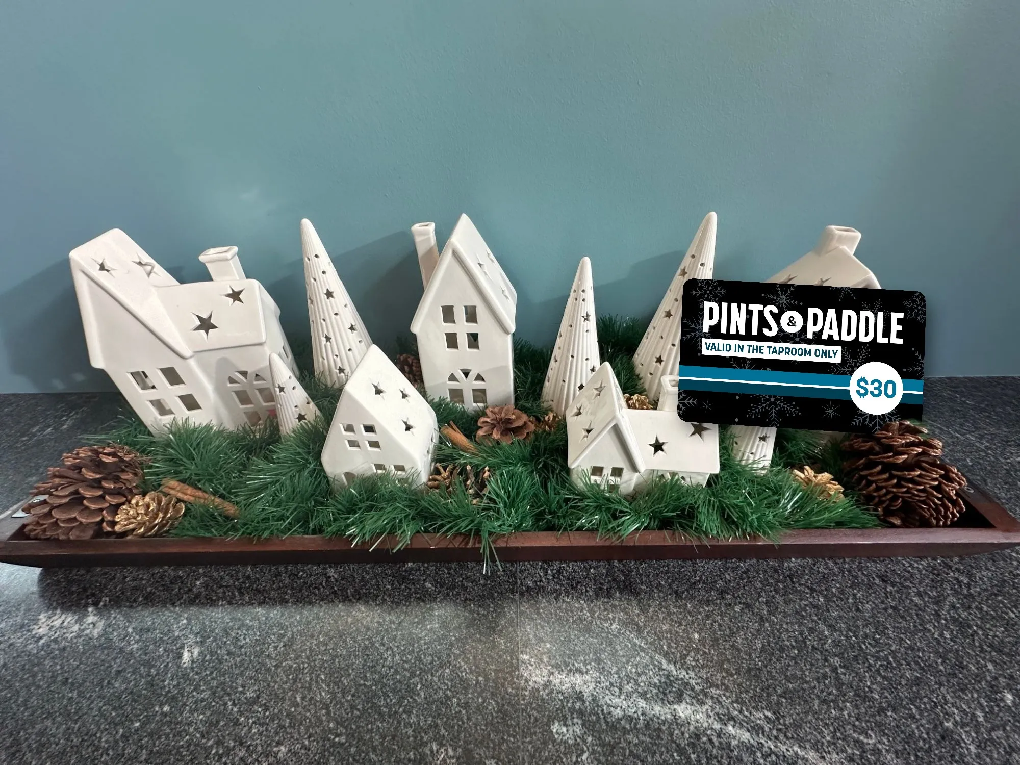 A $30 holiday bonus card propped in the greenery of a white ceramic holiday village, houses and trees with star cut-outs on a wooden tray