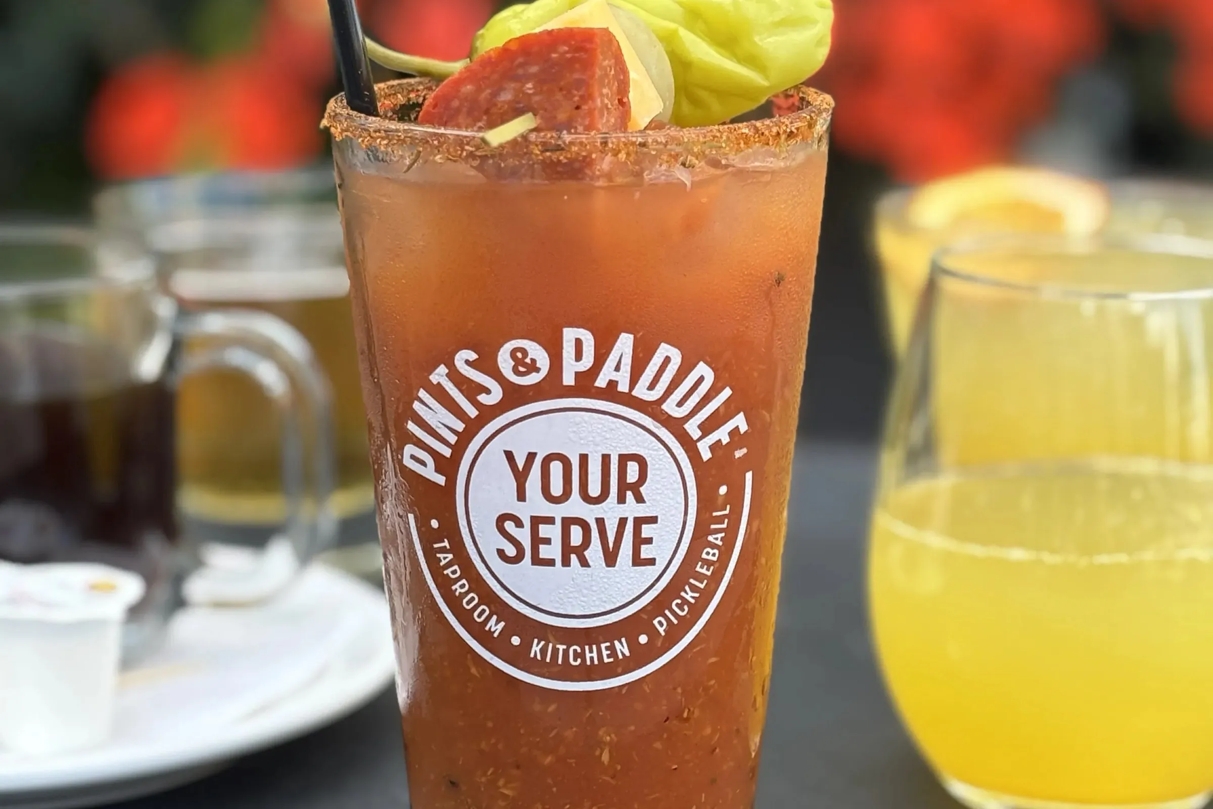 A bloody mary in a Pints & Paddle Your Serve pint glass, garnished with pepperoni, pickles and a pepperoncini, beside a mimosa and a mug of coffee on a patio tray