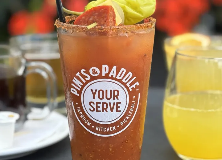 A bloody mary in a Pints & Paddle Your Serve pint glass, garnished with pepperoni, pickles and a pepperoncini, beside a mimosa and a mug of coffee on a patio tray