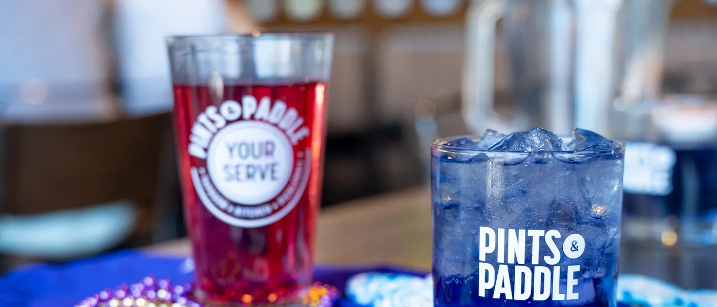 A Your Serve pint of red beer and a Grape Ape on a purple Paddy jersey with beads, the TVs over the bar blurred behind