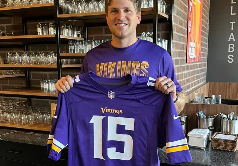 A smiling raffle winner in a Vikings tee holds up a purple number 15 jersey in front of the glassware shelves and the pull-tabs counter