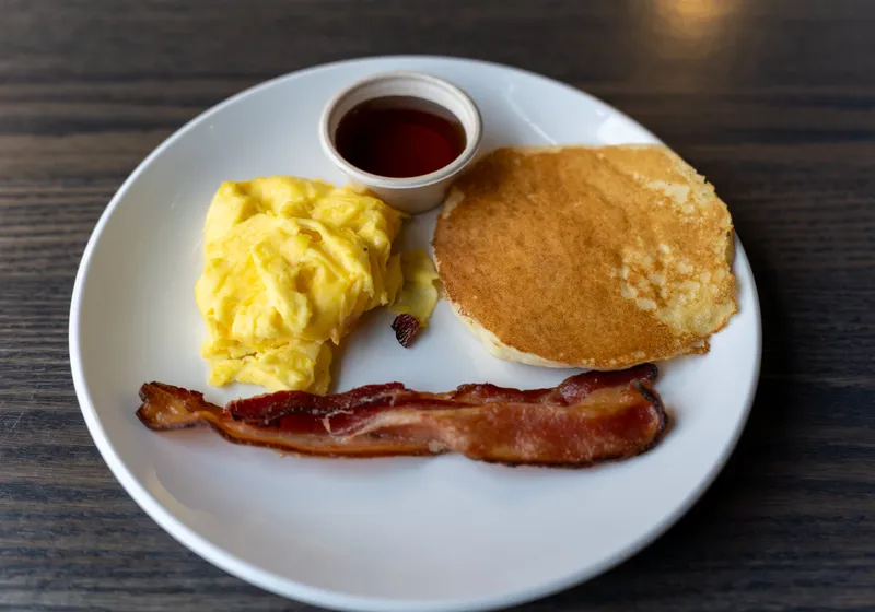 A kids breakfast plate with scrambled eggs, a small pancake, a strip of bacon and a cup of syrup