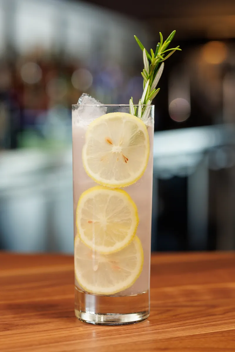 A tall glass of pale lavender-pink lemonade with three stacked lemon wheels, ice and a rosemary sprig