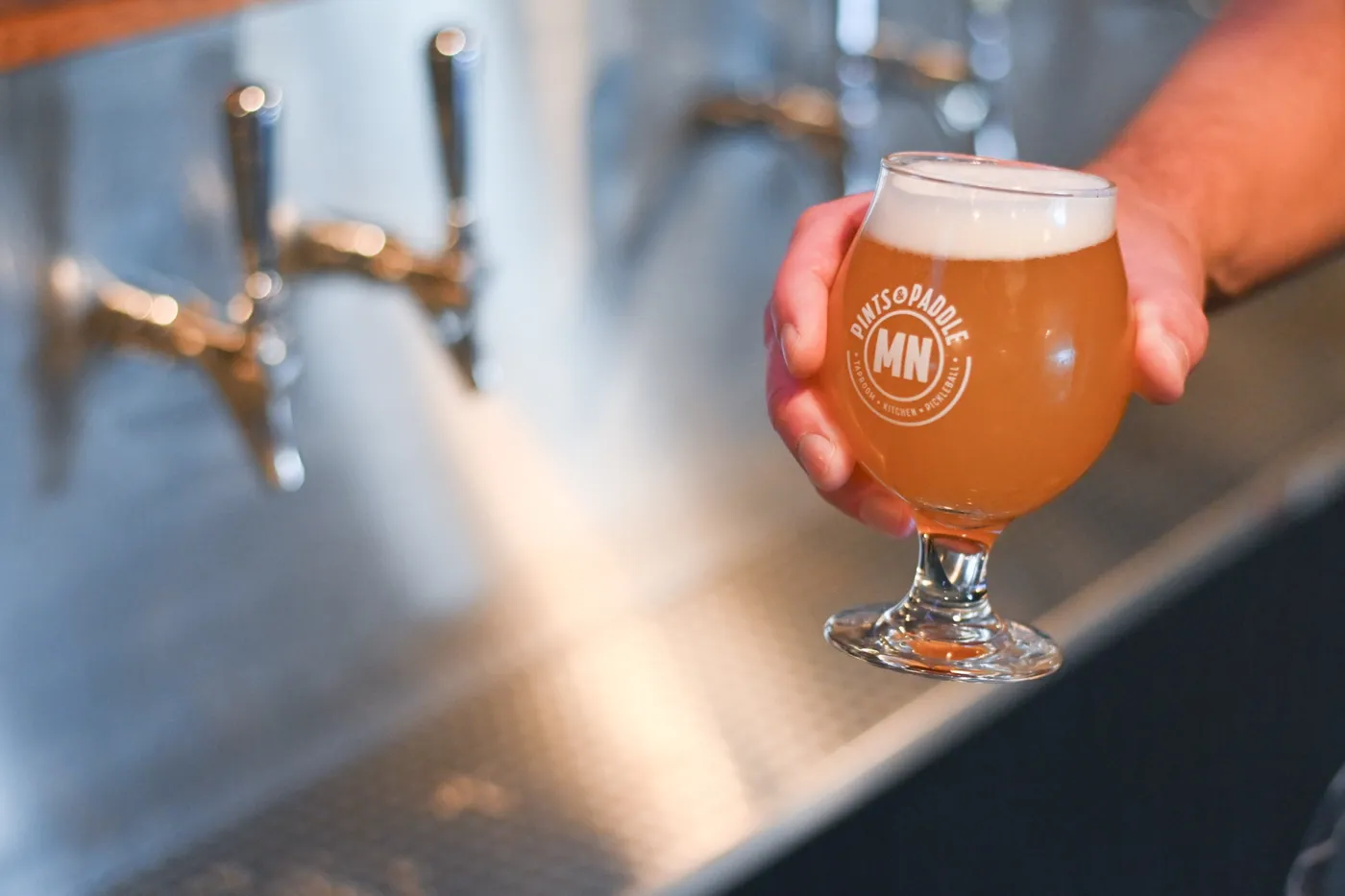 A hand holds out a full tulip glass of hazy golden beer with a thick white head, the Pints & Paddle MN roundel facing the camera, in front of the chrome faucets of the self-serve tap wall