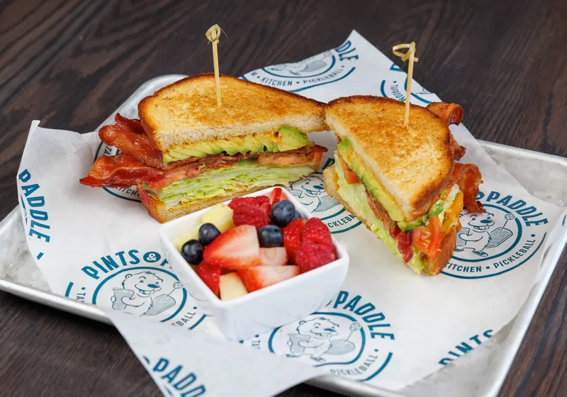 An avocado BLT cut in half showing bacon, avocado, lettuce and tomato on toasted bread, with a cup of fresh fruit