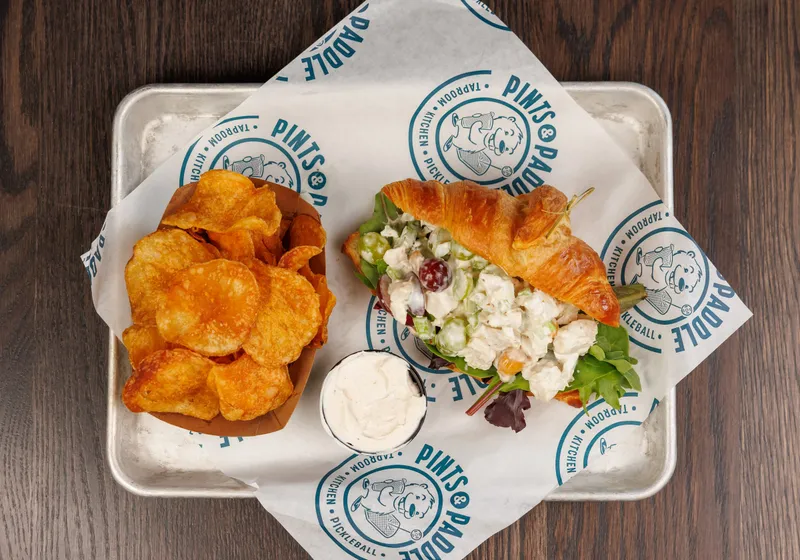 A chicken salad croissant from above with a boat of house kettle chips and a cup of dip on branded paper
