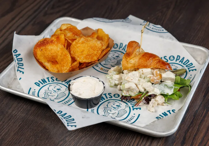 Half a chicken salad croissant with mixed greens, a boat of kettle chips and a cup of dip on Pints & Paddle paper
