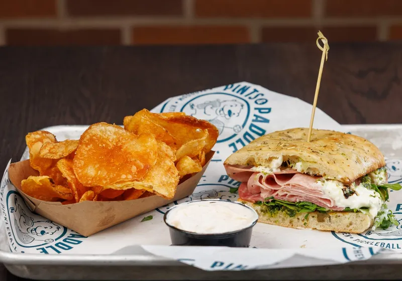 Half a Toscana sandwich on focaccia with mortadella, salami and burrata showing, kettle chips and a cup of dip, brick wall behind