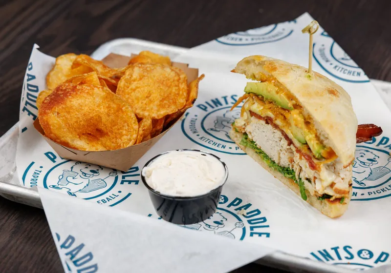 Half a turkey avocado sandwich on focaccia with a boat of kettle chips and a cup of dip on a Pints & Paddle tray