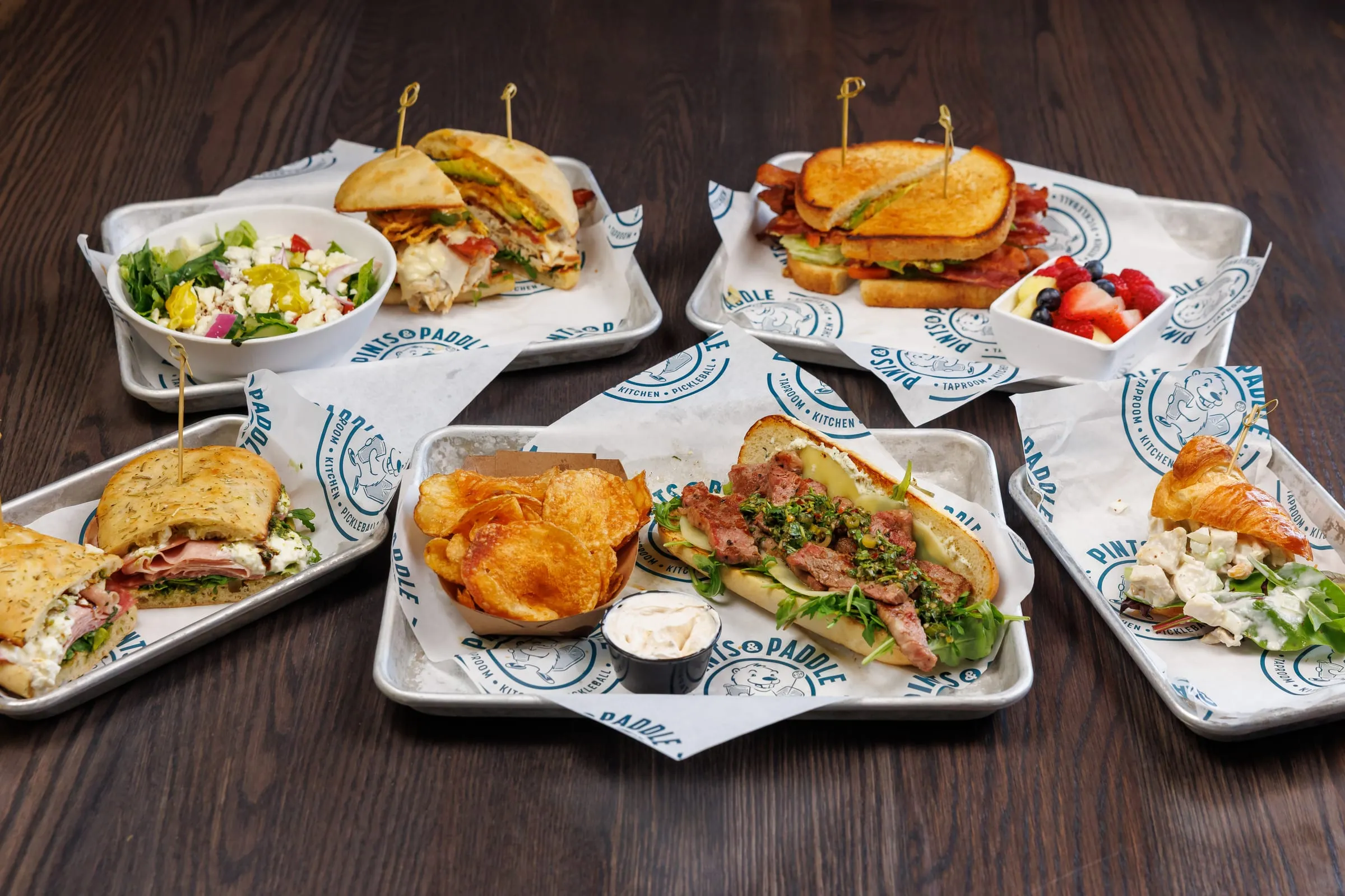 The lunch spread at an angle: sandwiches, kettle chips, a side salad and a fruit cup on Pints & Paddle trays