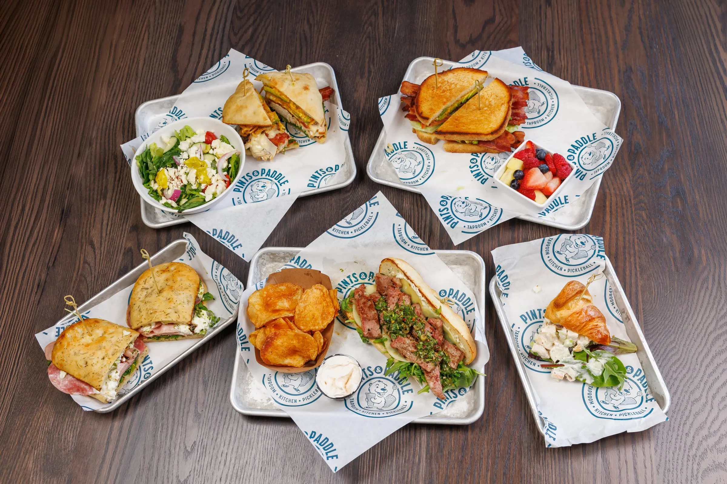 The weekday lunch spread from above: five sandwiches on Pints & Paddle trays with kettle chips, a side salad and a fruit cup