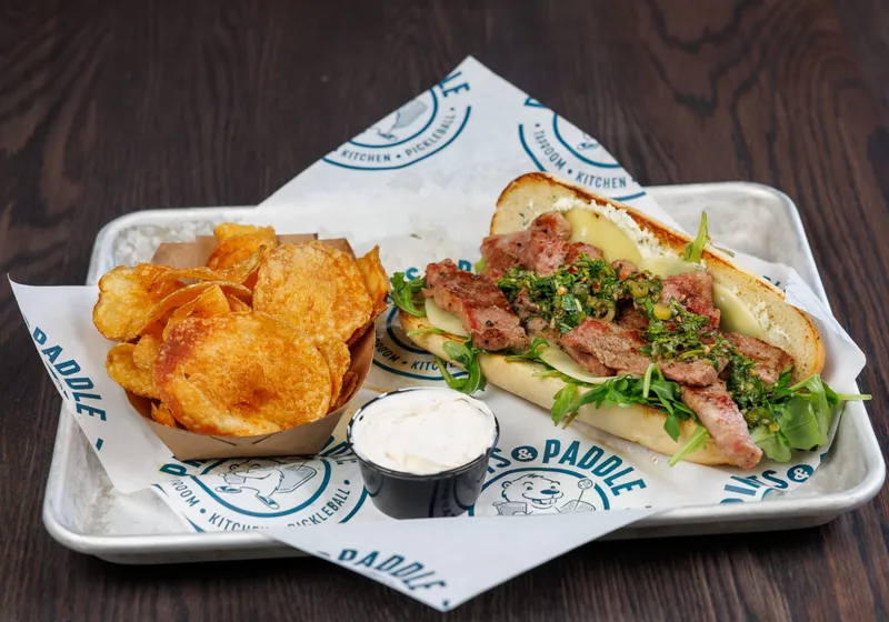 A steak sandwich on a hoagie roll piled with sliced steak, arugula and chimichurri, with kettle chips and a cup of dip