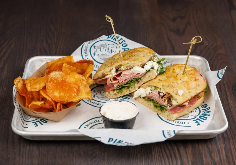 A Toscana sandwich cut in half on focaccia, mortadella, salami and burrata showing, with kettle chips and a cup of dip