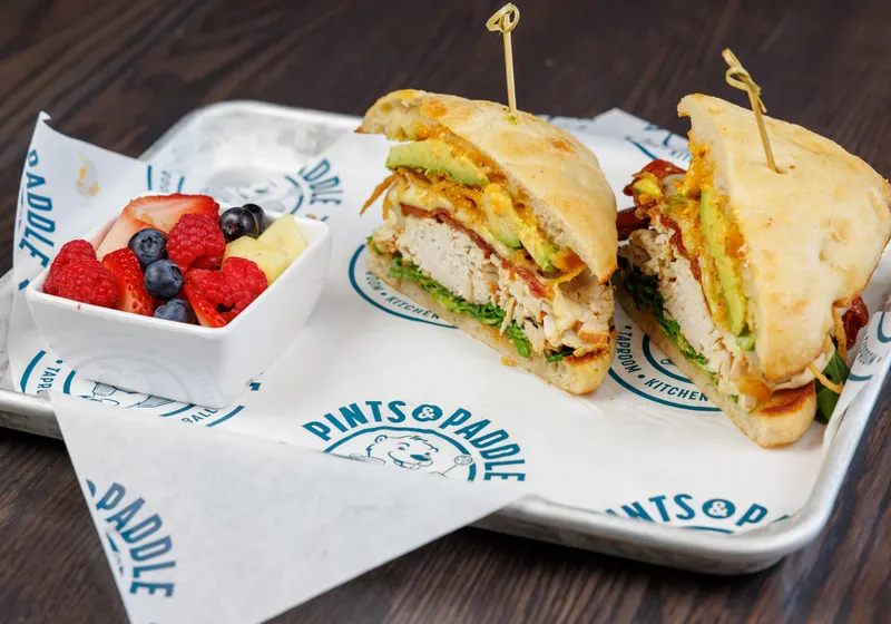 A turkey avocado sandwich cut in half on focaccia, avocado and crispy onions showing, with a cup of fresh fruit