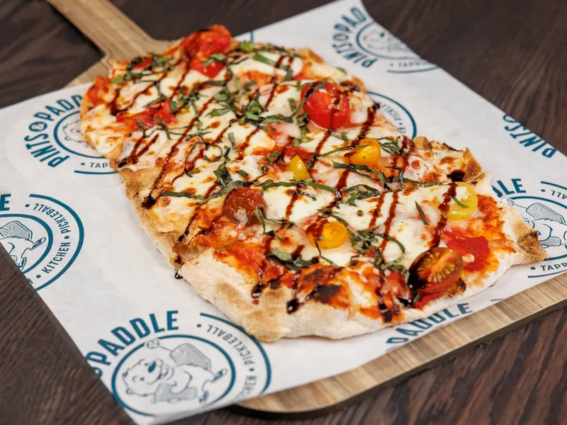 A thin-crust Margherita pizza with cherry tomatoes, fresh mozzarella, basil and balsamic drizzle on a wooden pizza peel