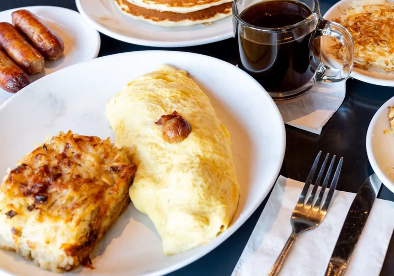 A sausage omelette beside a square of cheesy hash browns, with a mug of coffee, sausage links and pancakes behind
