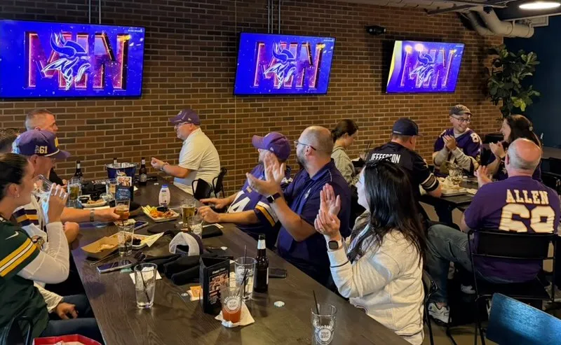 Fans in Vikings jerseys clapping at long tables on the mezzanine, the MIN logo on all three screens along the brick wall