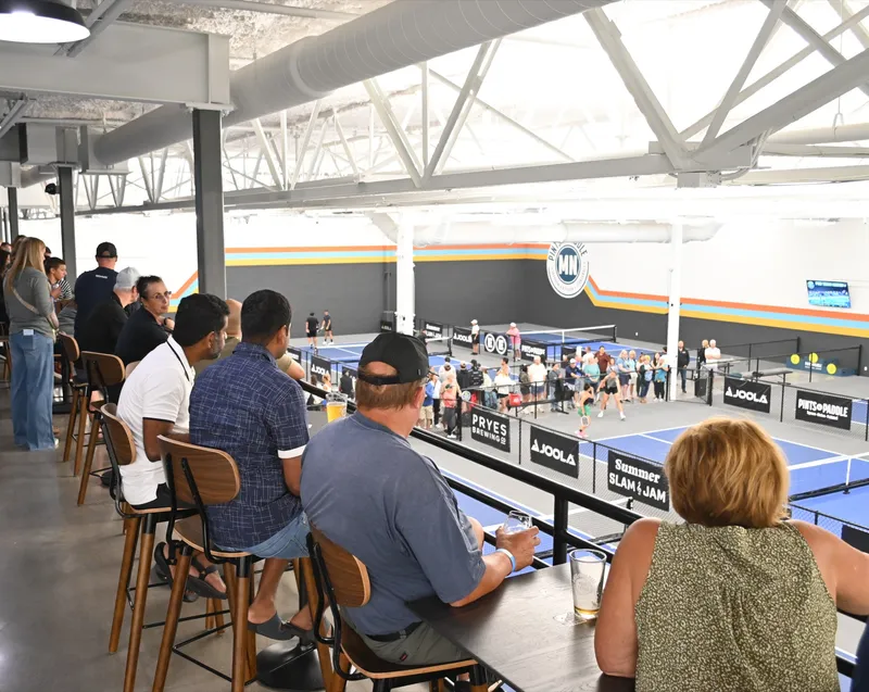 Guests on stools along the mezzanine drink rail with beers, watching a tournament match on the courts below